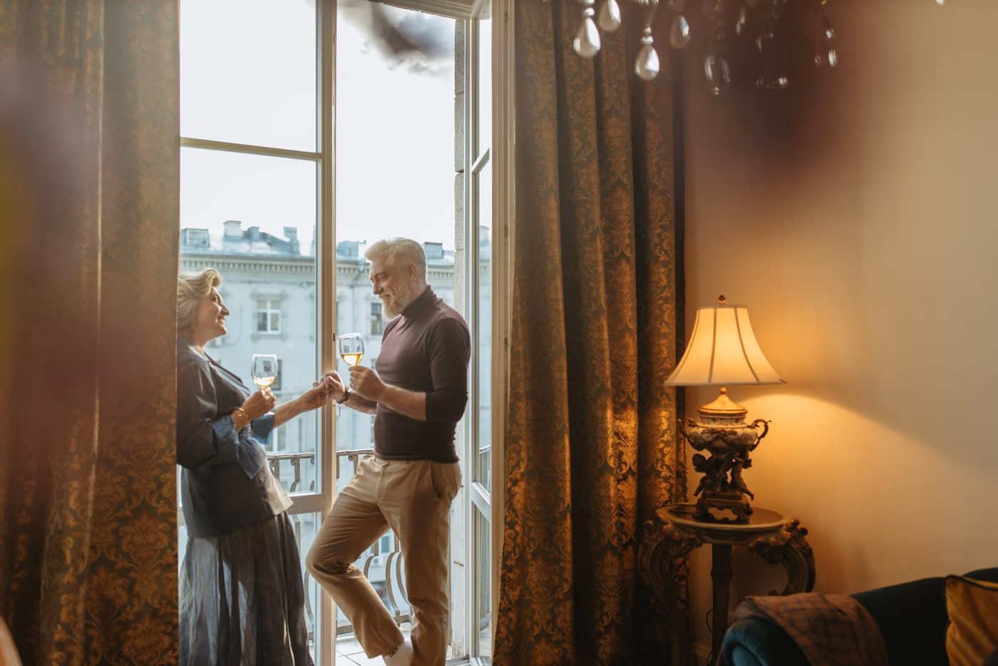 A mature couple sharing wine beside tall windows in a warmly lit, ornate room